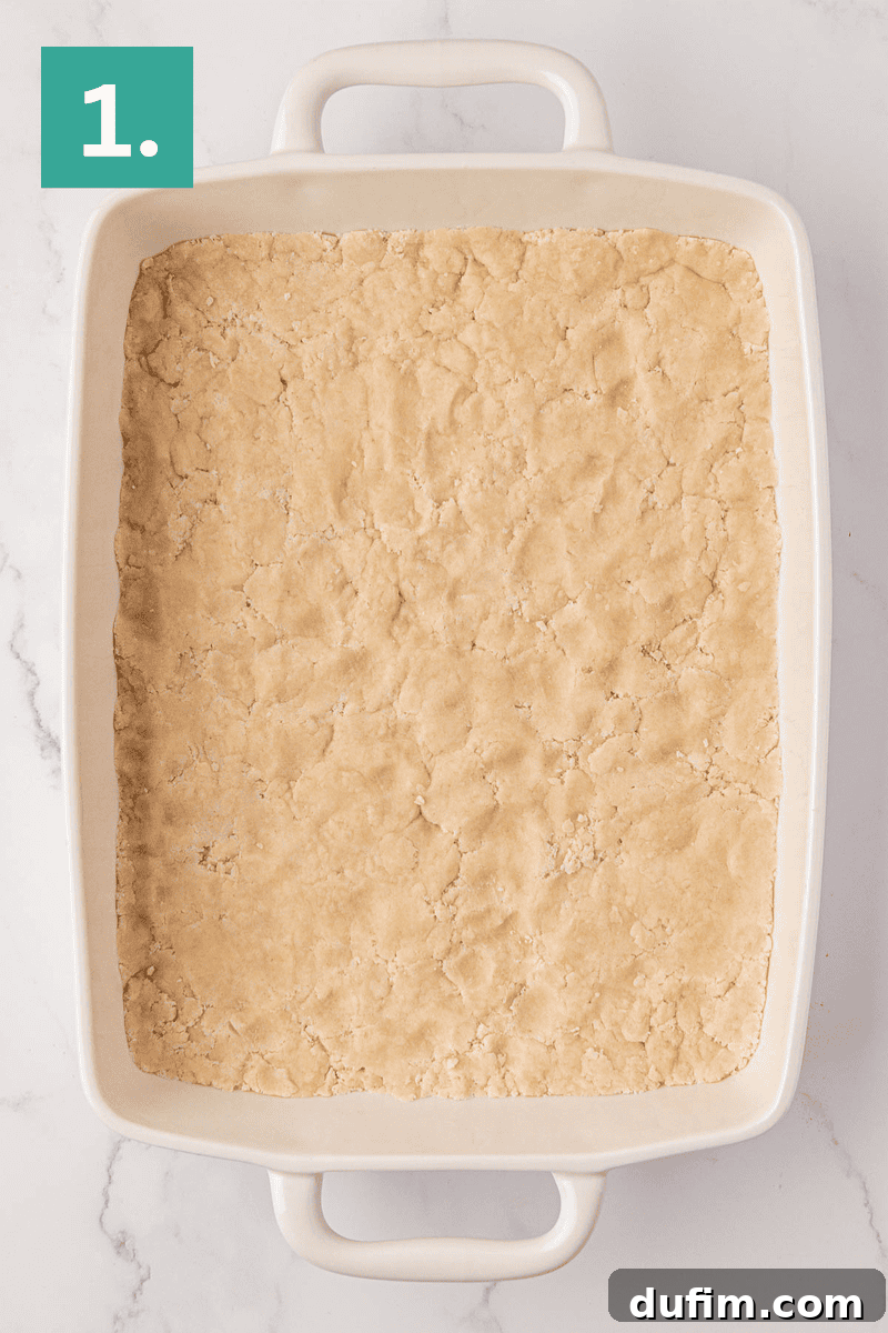 A rectangular white baking dish is meticulously filled with an even, golden-brown layer of unbaked, buttery dough, ready for the next step. A vibrant green square in the top left corner prominently displays the number “1,” indicating the first step in the recipe.