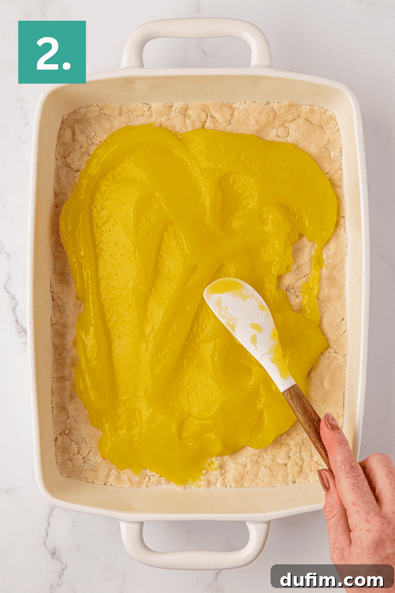 A person uses a pristine white spatula to smoothly spread a generous layer of bright yellow lemon filling over a perfectly baked, golden crust within a rectangular white baking dish. A green box with the number 2 in the top left corner visually marks this as the second step.