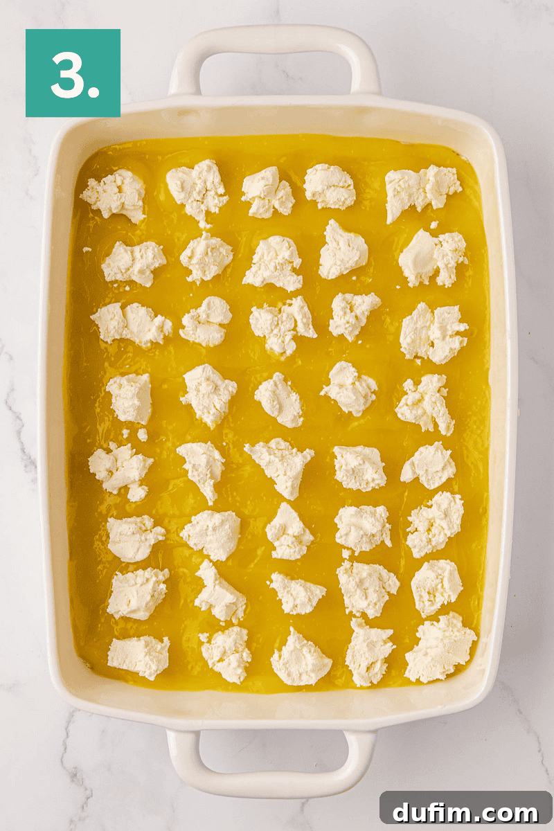 A crisp white baking dish is layered with a vibrant yellow filling, generously topped with an array of evenly spaced, soft white dollops of cream cheese. A prominent green box in the top left corner highlights the number 3, indicating this step in the baking process.