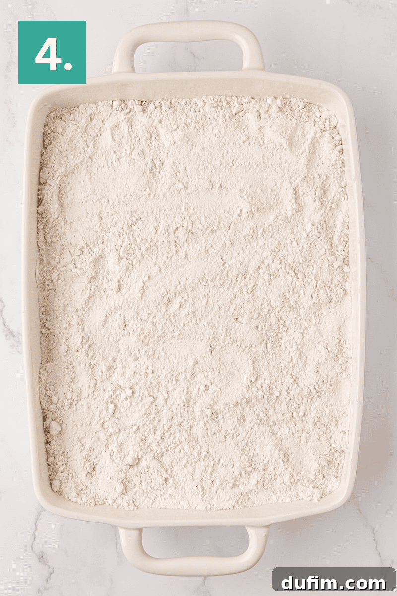 A rectangular white baking dish, filled with a smooth, even layer of cake mix, rests on a pristine white marble surface, ready for the next stage of baking. A distinct green box positioned in the top left corner displays the number 4, signifying the progression of the recipe.