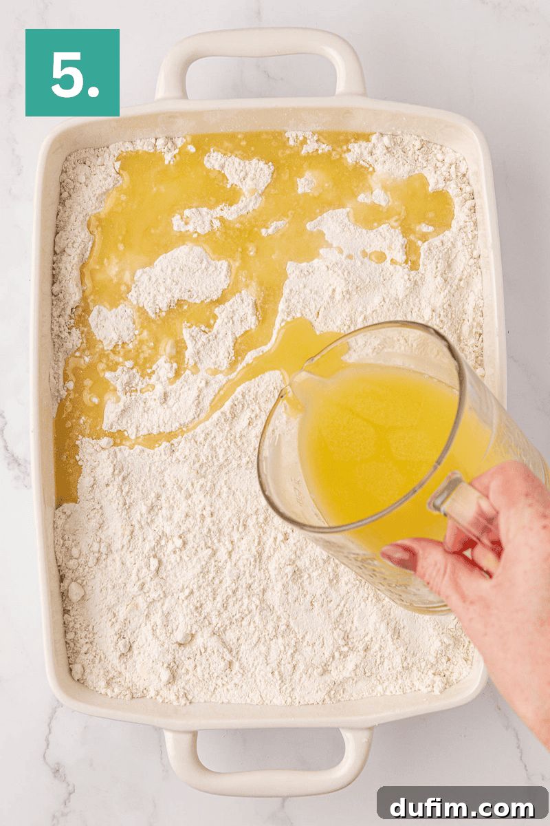A skilled hand carefully pours a stream of golden melted butter from a clear measuring cup onto a pristine, even layer of flour in a white rectangular baking dish. In the upper left corner, a distinct green box with the number 5 clearly indicates the current step in the recipe.