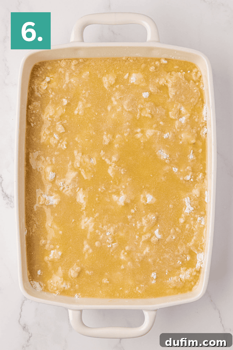 A pristine white baking dish contains an evenly distributed layer of dry cake mix, lightly moistened with drizzled butter, creating a textured surface ready for baking. The number 6 in a green box on the top left corner indicates the final preparation step.