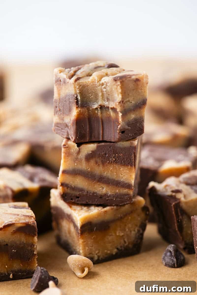 Decadent Chocolate Peanut Butter Fudge 2 Three pieces of marbled chocolate and peanut butter fudge are stacked on top of each other, with more fudge pieces and chocolate chips scattered in the background on a brown surface.