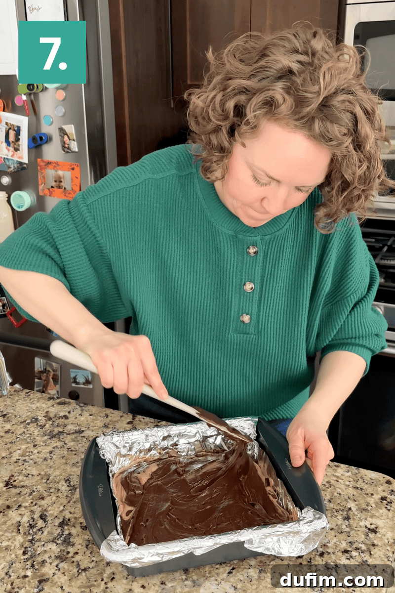 Decadent Chocolate Peanut Butter Fudge 12 A woman in a green sweater spreads chocolate over a foil-lined pan on a granite kitchen counter. Step 7 is indicated in the top left corner. Refrigerator and cabinets are visible in the background.