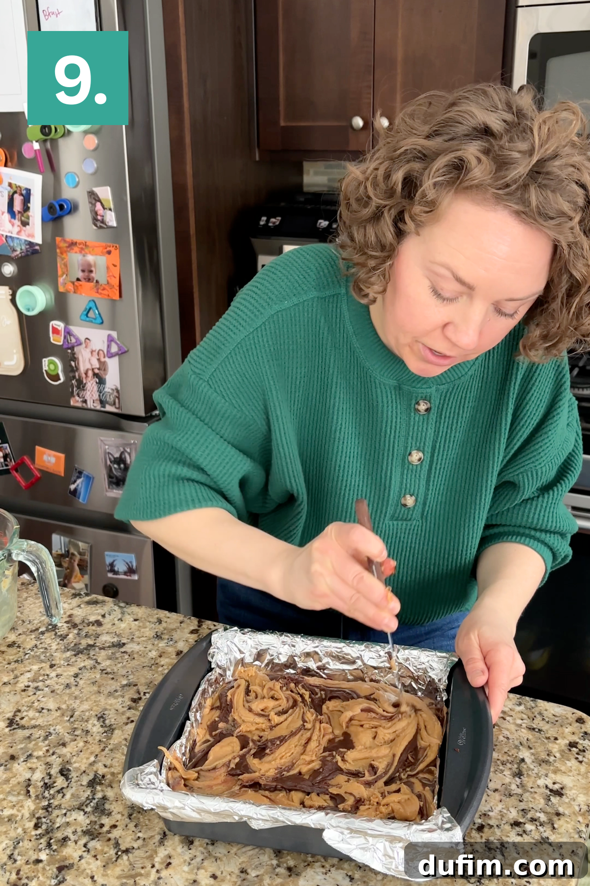 Decadent Chocolate Peanut Butter Fudge 14 A woman in a green sweater swirls batter in a foil-lined baking pan on a kitchen counter. A refrigerator with photos and magnets is in the background, and a number 9 appears in the top left corner.