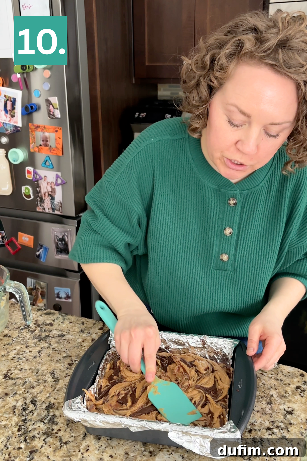 Decadent Chocolate Peanut Butter Fudge 15 A woman in a green shirt spreads marbled batter in a foil-lined square baking pan with a spatula on a kitchen counter. A fridge with magnets and photos is in the background. A green box with 10. is in the top left corner.