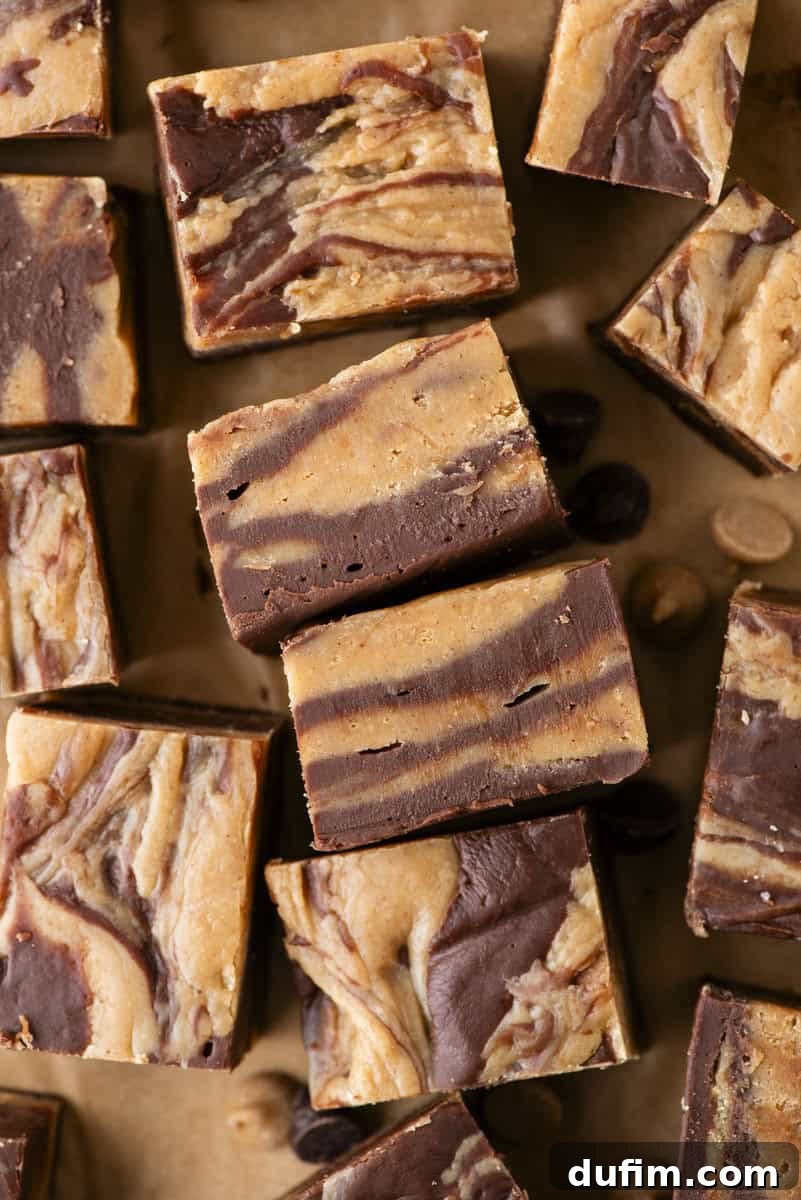 Decadent Chocolate Peanut Butter Fudge 5 Squares of marbled chocolate and peanut butter fudge are arranged on a brown surface. The fudge has swirled layers of dark brown chocolate and light brown peanut butter. A few chocolate and peanut butter chips are scattered nearby.