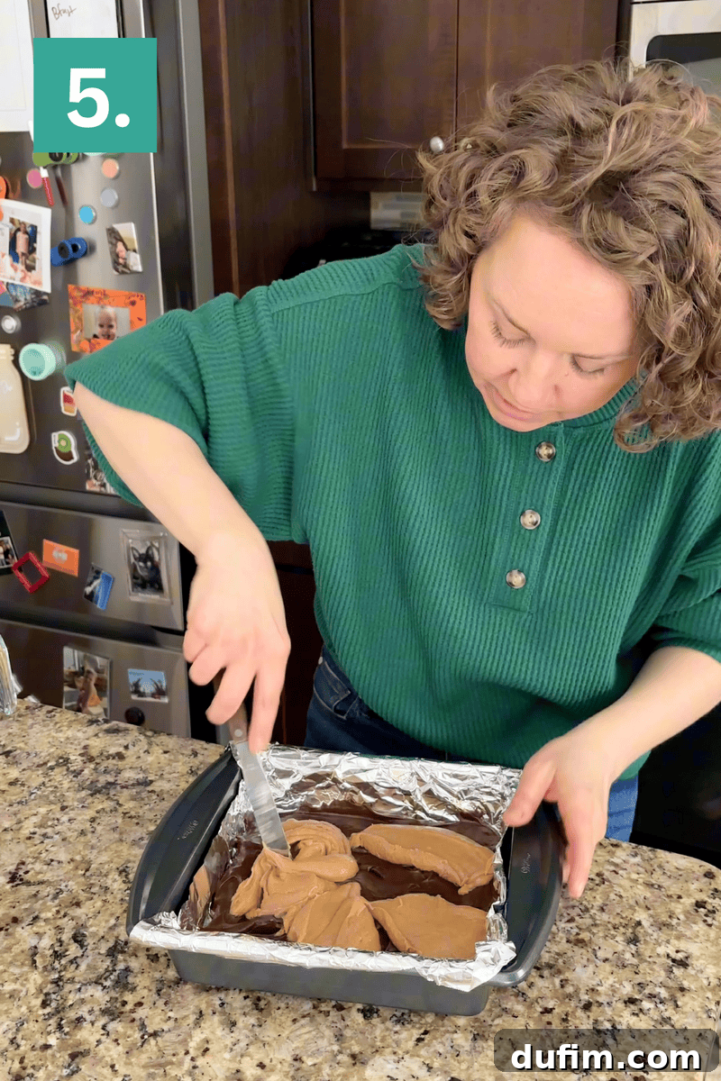 Decadent Chocolate Peanut Butter Fudge 10 A person in a green sweater spreads chocolate frosting on a dessert in a foil-lined baking pan on a granite countertop. A fridge with photos and magnets is in the background. A green box with the number 5 is in the top left corner.
