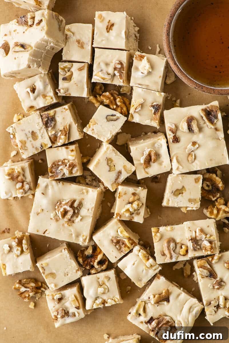 Squares of creamy maple walnut fudge topped with chopped walnuts are scattered on brown parchment paper next to a small cup of maple syrup.