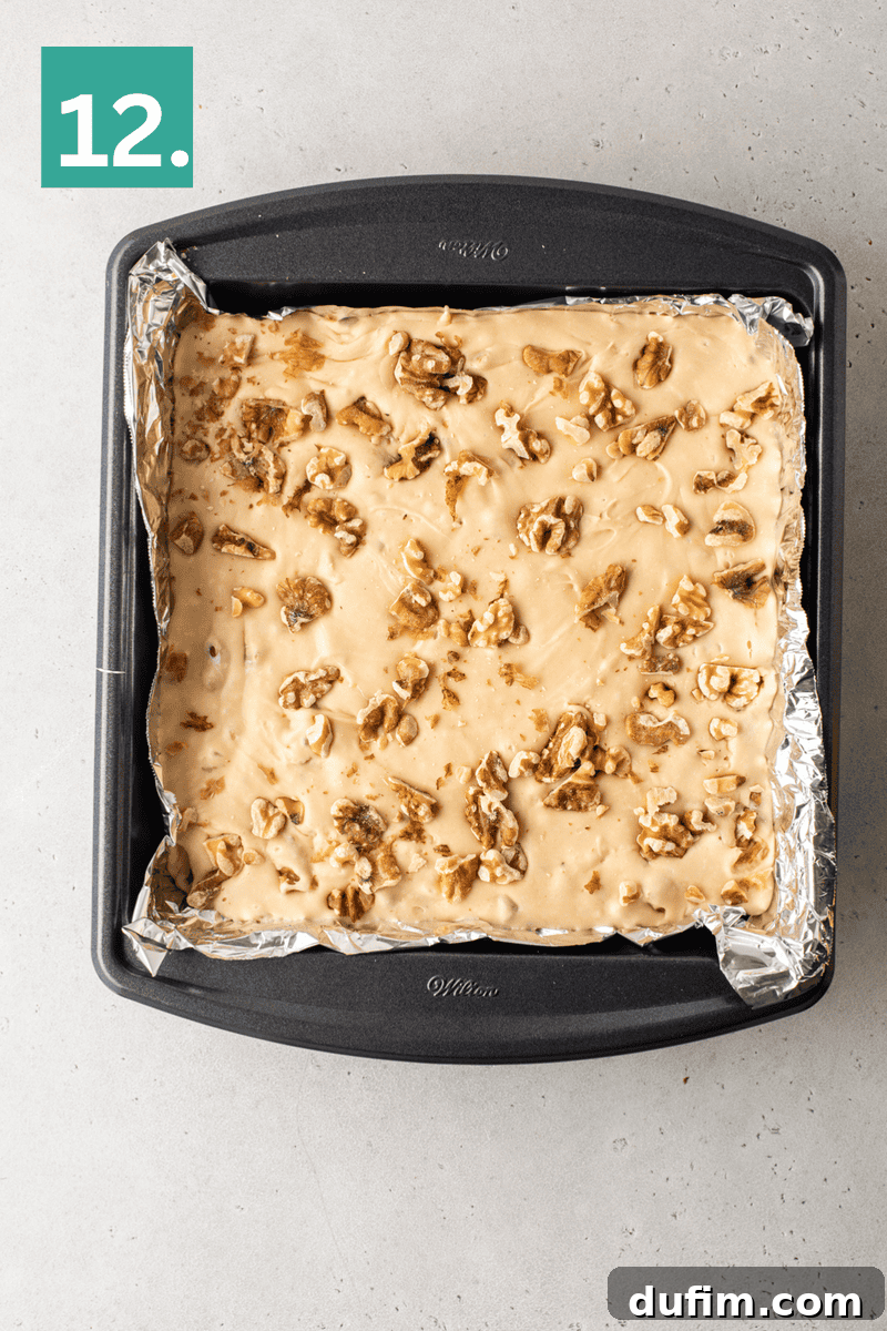 A square baking pan lined with foil holds a layer of beige fudge topped with chopped walnuts. A green box with the number 12 is in the top left corner. The pan rests on a light gray surface.