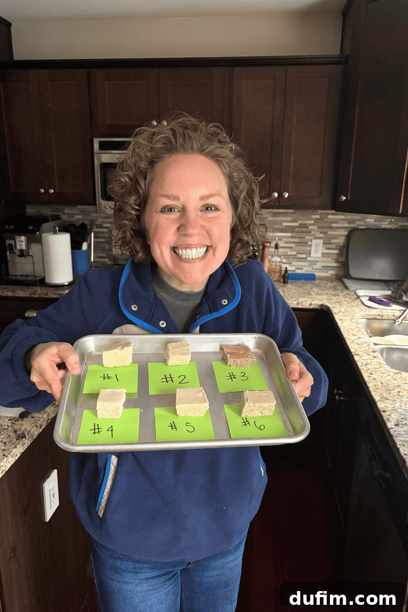 A smiling person with curly hair holds a baking tray with six pieces of dessert, each labeled with a number on green paper, standing in a kitchen with dark cabinets and a granite countertop.