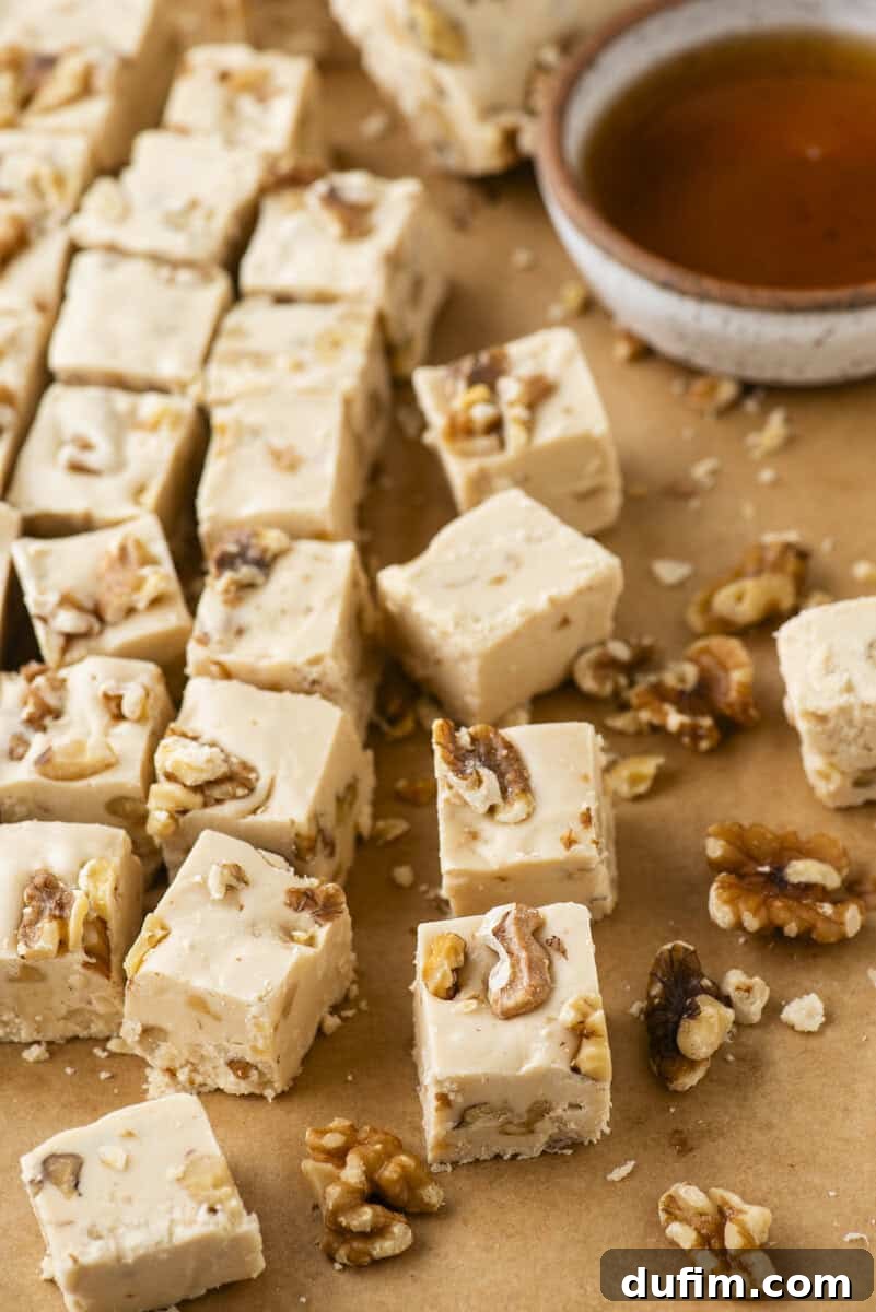 Cubes of maple walnut fudge are arranged on brown parchment paper, surrounded by scattered walnut pieces. A small bowl of maple syrup sits nearby.