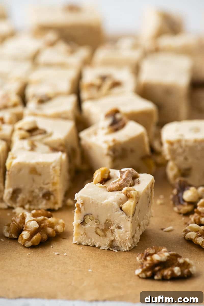 Close-up of creamy fudge squares with pieces of walnuts on top and inside, arranged on brown parchment paper. Some walnut halves are scattered around the fudge pieces.