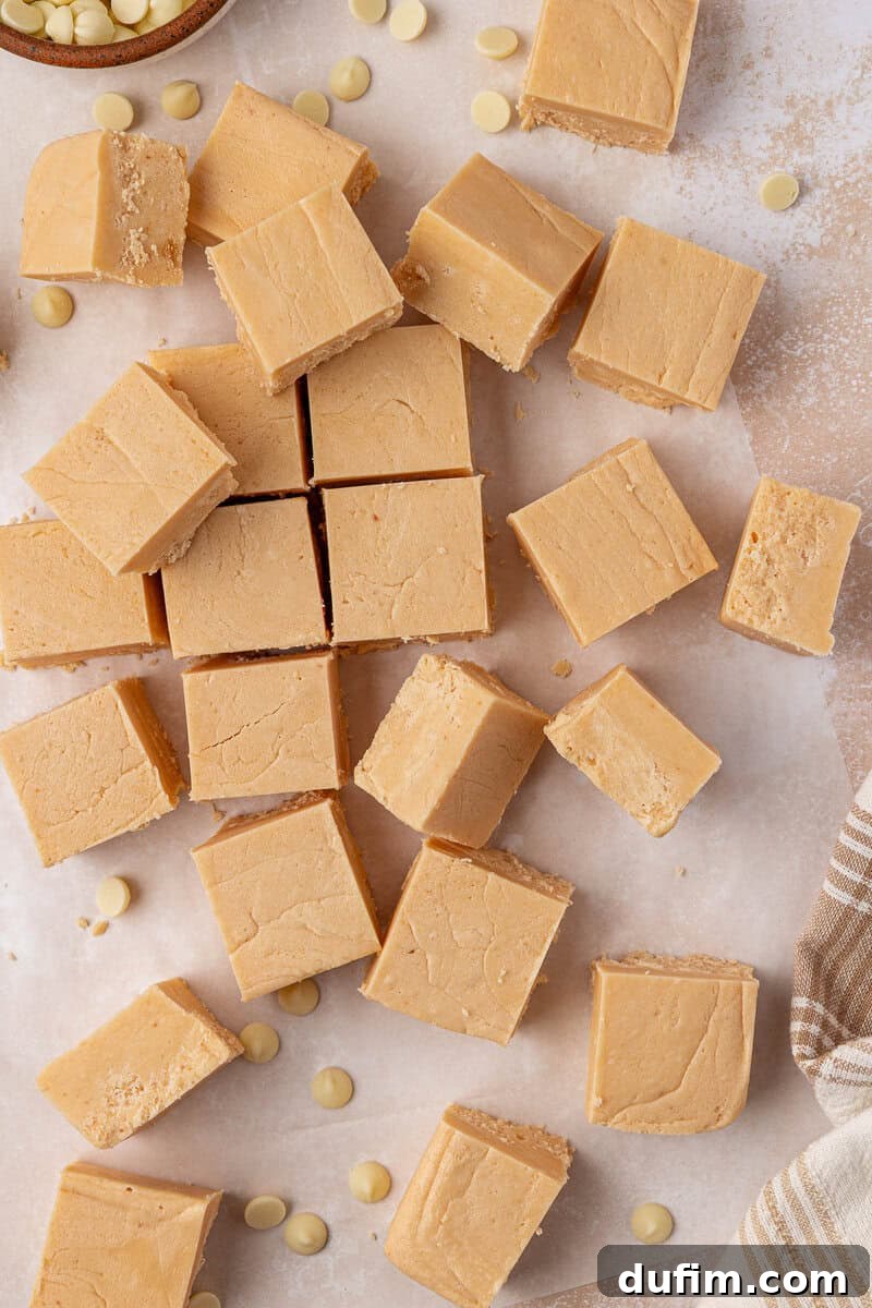 Guaranteed Perfect Vanilla Fudge 2 Squares of creamy vanilla fudge are arranged on parchment paper, with a few white chocolate chips scattered around. A striped cloth is partially visible in the corner.