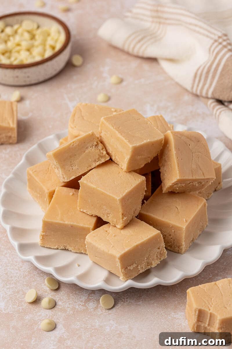 Guaranteed Perfect Vanilla Fudge 11 A plate stacked with creamy beige fudge squares, surrounded by scattered white chocolate chips. A bowl of more chocolate chips and a striped cloth are in the background.