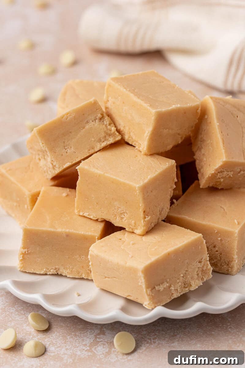 Guaranteed Perfect Vanilla Fudge 13 A stack of creamy, light brown fudge squares arranged on a white, scalloped-edged plate with a few white chocolate chips scattered nearby.