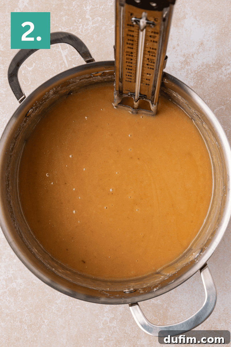 Guaranteed Perfect Vanilla Fudge 5 A saucepan filled with light brown caramel mixture sits on a countertop, with a candy thermometer clipped to the side. A green box in the corner displays the number 2.
