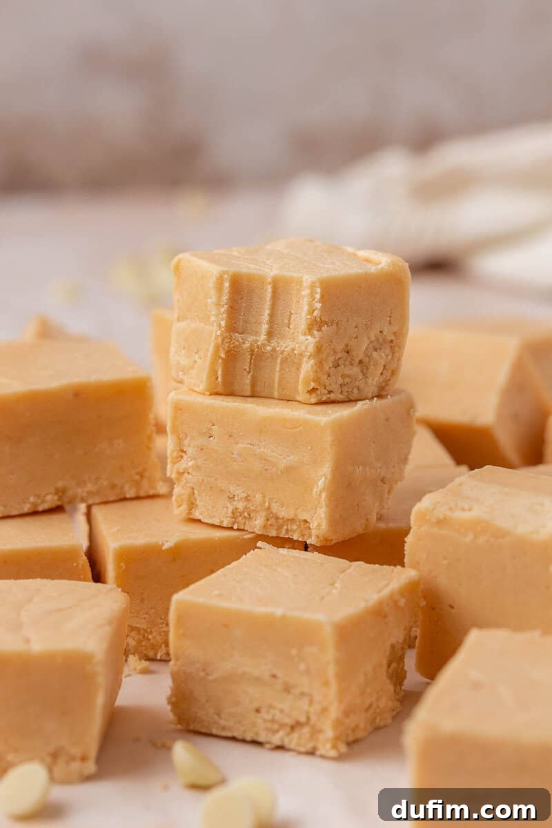 Guaranteed Perfect Vanilla Fudge 10 A stack of creamy, light brown fudge squares sits on a surface, with the top piece showing bite marks. More fudge pieces and white chocolate chips are scattered around.