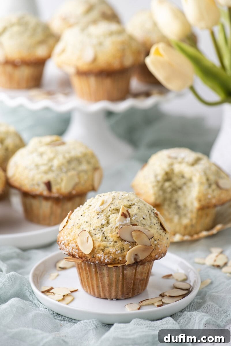 Irresistible Almond Poppy Seed Muffins 2 A perfectly baked almond poppy seed muffin, garnished with sliced almonds, rests on a small white plate. In the blurred background, more muffins and a vibrant bouquet of yellow tulips create an inviting scene.