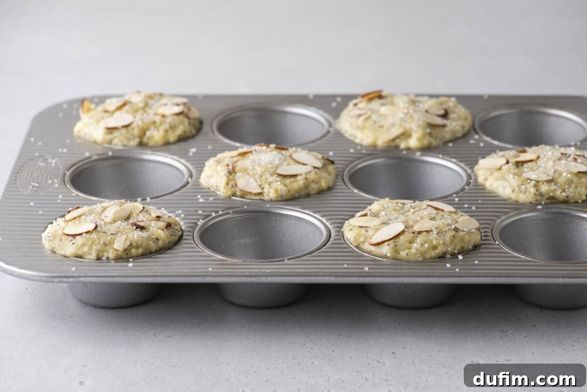 Irresistible Almond Poppy Seed Muffins 11 A metal muffin tin is ready for baking, with six muffin wells filled generously with thick, domed almond poppy seed batter, each topped with sliced almonds and coarse sanding sugar. The alternating empty wells promote a better rise.