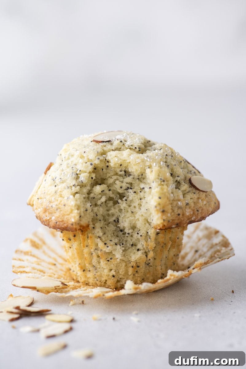 Irresistible Almond Poppy Seed Muffins 14 A close-up shot of a delectable almond poppy seed muffin, with a bite already taken, revealing its fluffy and moist interior. Sliced almonds and scattered crumbs lie nearby on a simple light surface, inviting another taste.