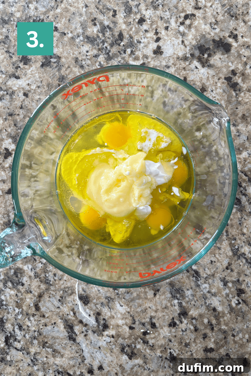 Irresistible Almond Poppy Seed Muffins 7 A clear glass Pyrex measuring cup holds the wet ingredients: eggs, buttermilk, sour cream, oil, melted butter, and almond extract, glistening on a granite countertop.