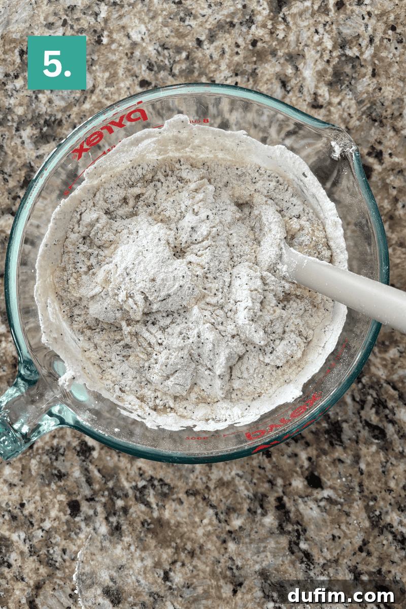 Irresistible Almond Poppy Seed Muffins 9 The process of gradually mixing dry ingredients into the wet mixture in a clear glass bowl, showing the flour being incorporated into the liquids.