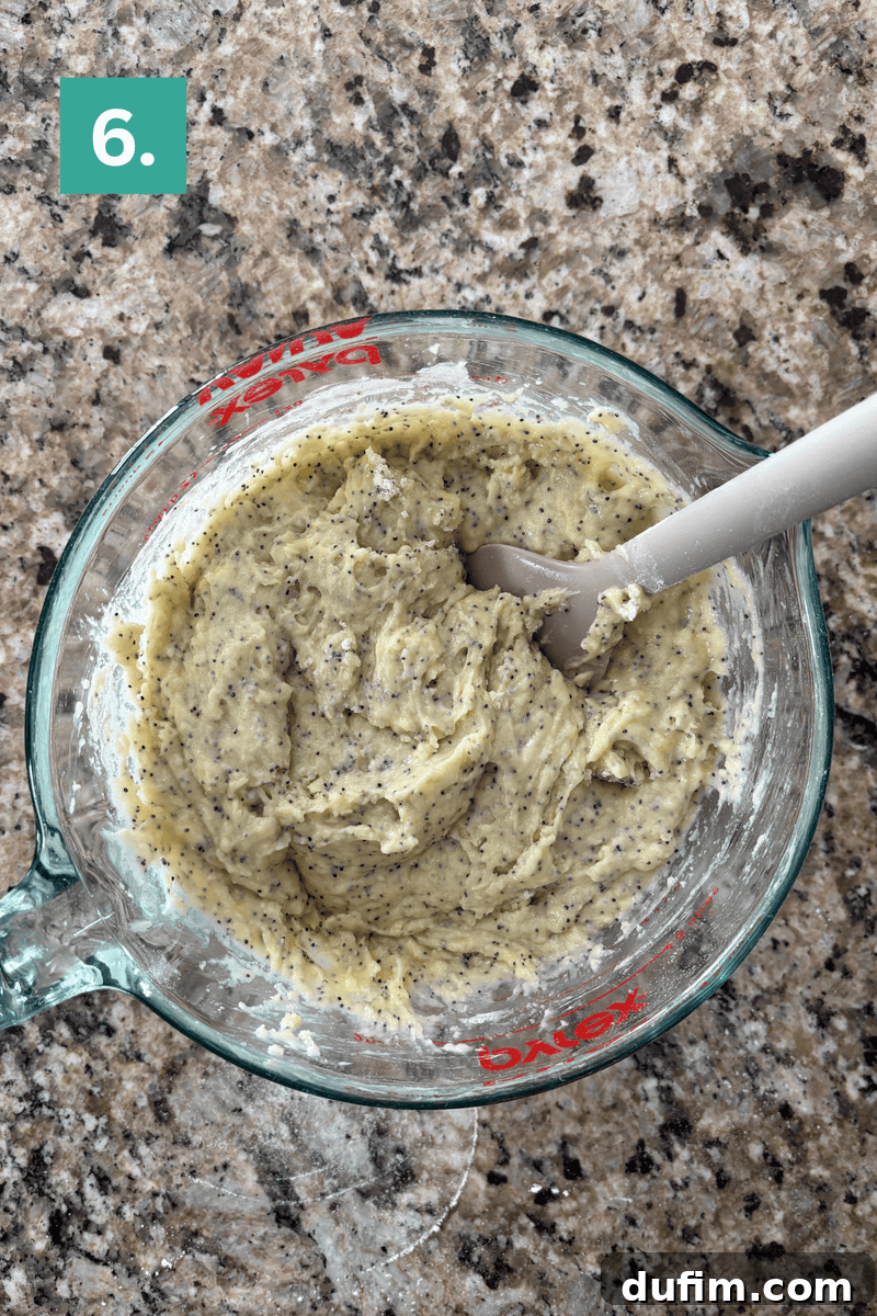 Irresistible Almond Poppy Seed Muffins 10 A thick, rich almond poppy seed muffin batter being gently folded with a gray spatula in a glass mixing bowl, revealing its dense yet smooth consistency with visible poppy seeds.
