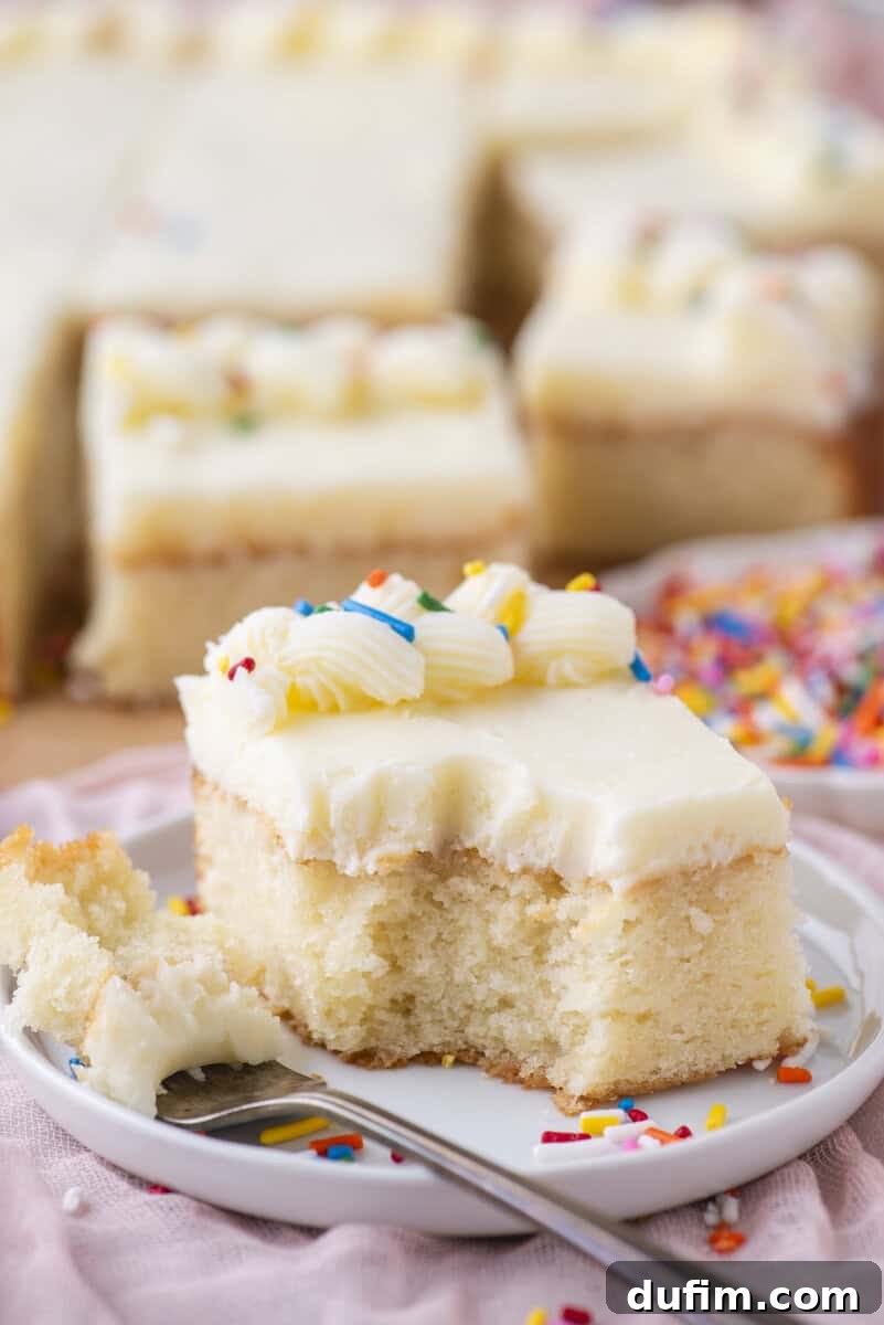 A slice of vanilla cake with white frosting and colorful sprinkles sits on a white plate, with a bite taken out and a fork nearby. More cake pieces and sprinkles are blurred in the background.