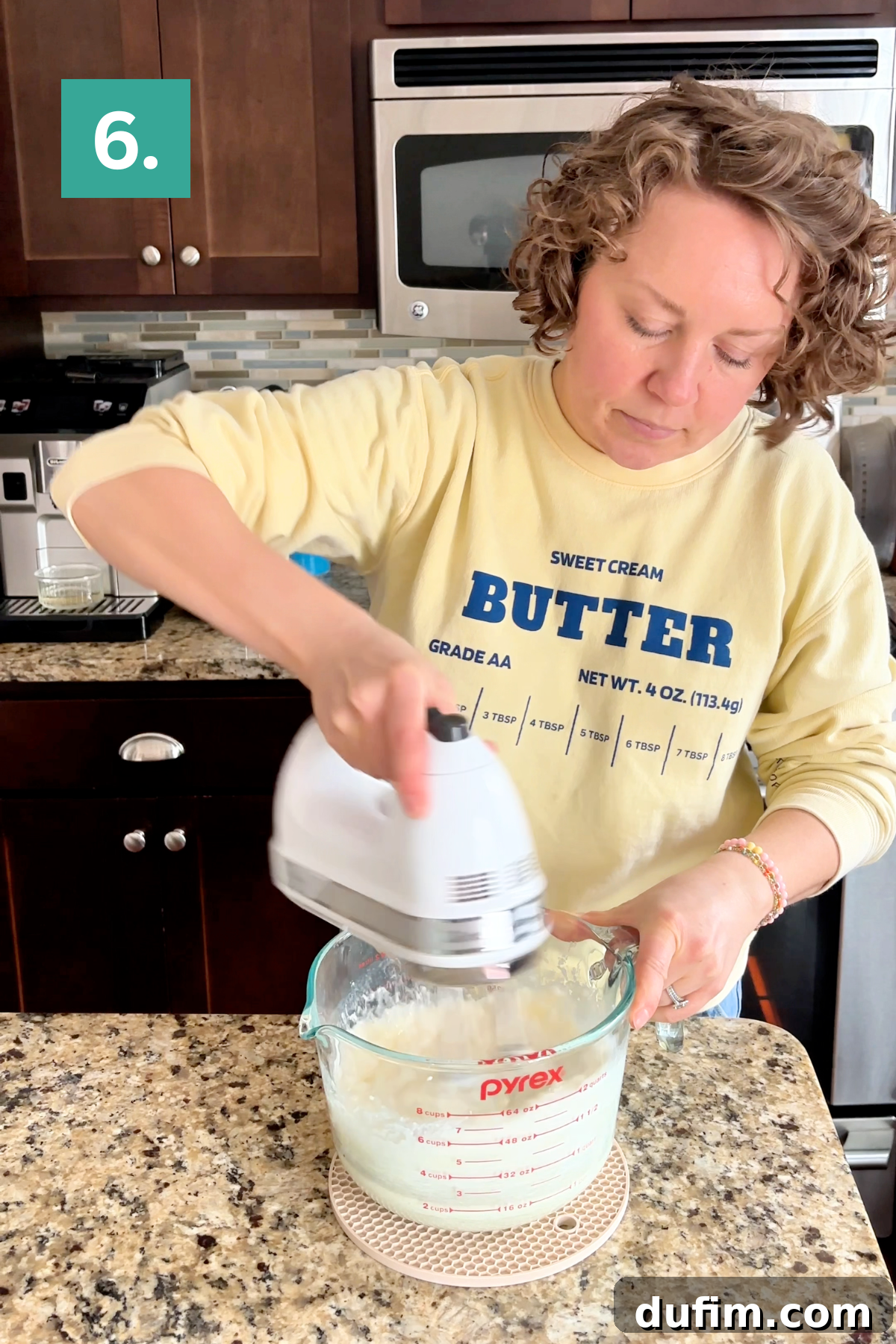 A woman wearing a yellow Butter sweatshirt uses a handheld mixer to blend ingredients in a Pyrex measuring cup on a kitchen counter. A teal box with the number 6 is in the corner.