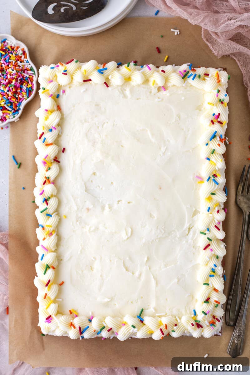 A rectangular sheet cake with white frosting, decorated with a piped border and colorful rainbow sprinkles, sits on brown parchment paper beside a small dish of extra sprinkles, a cake server, plates, and forks.