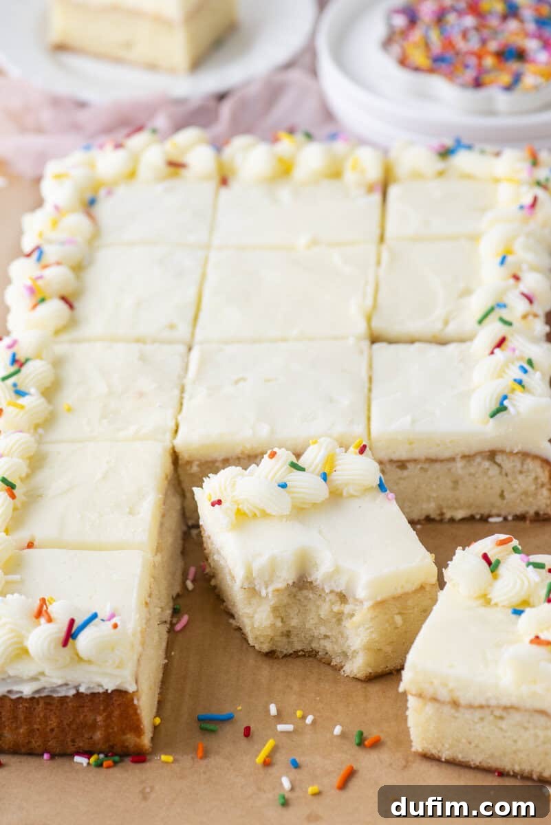 A rectangular white frosted sheet cake, cut into squares, with piped frosting and colorful sprinkles on the edges. One piece is pulled out in front with a bite taken from it. Plates with sprinkles are in the background.