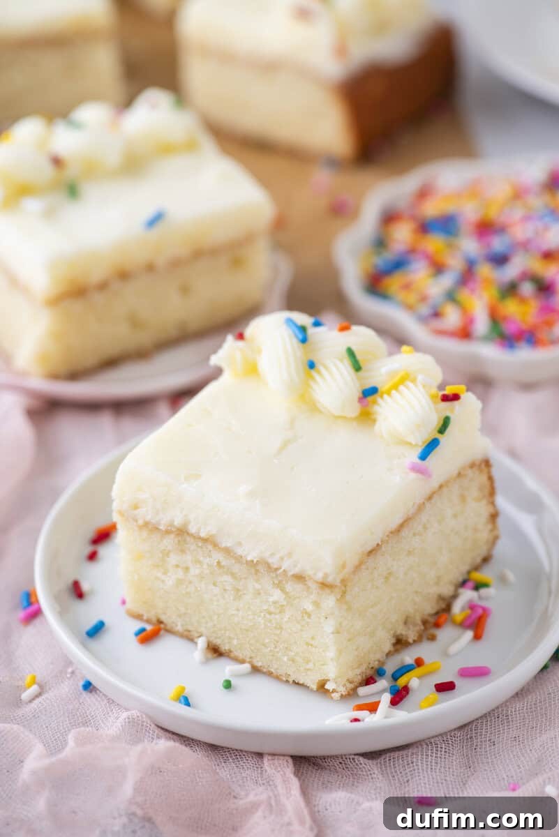 A square piece of vanilla cake with white frosting and colorful sprinkles sits on a white plate. More slices of cake and a small bowl of sprinkles are in the background.