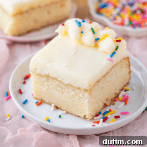 A square slice of vanilla cake with white frosting, decorated with piped frosting and colorful sprinkles, sits on a white plate with more sprinkles scattered around.