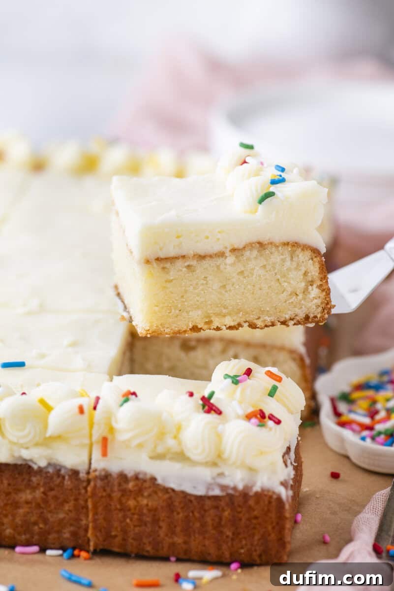 A slice of frosted vanilla sheet cake with colorful sprinkles is being lifted from the cake, which is decorated with piped white frosting and more sprinkles. A small dish of sprinkles sits nearby.