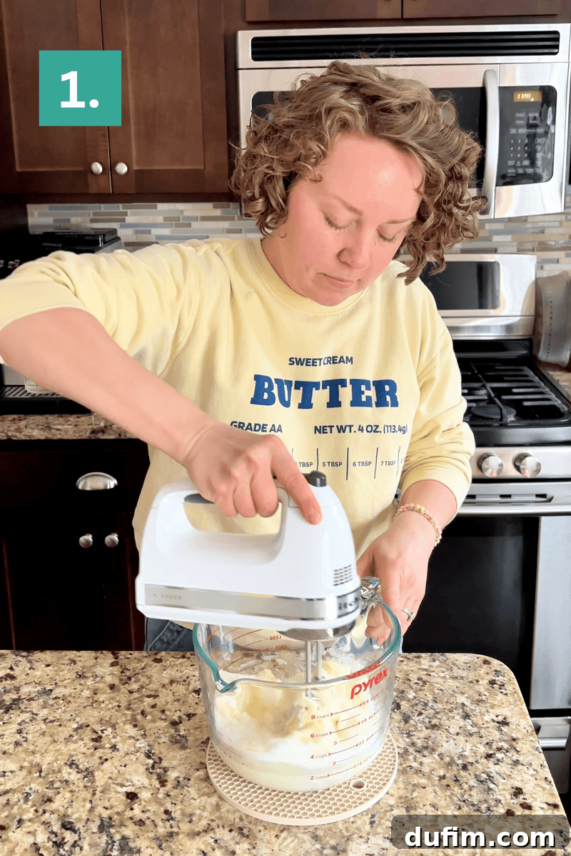 A person using a hand mixer to blend ingredients in a glass bowl on a granite kitchen counter. They are wearing a yellow BUTTER sweatshirt and standing in a kitchen with dark cabinets and stainless steel appliances.