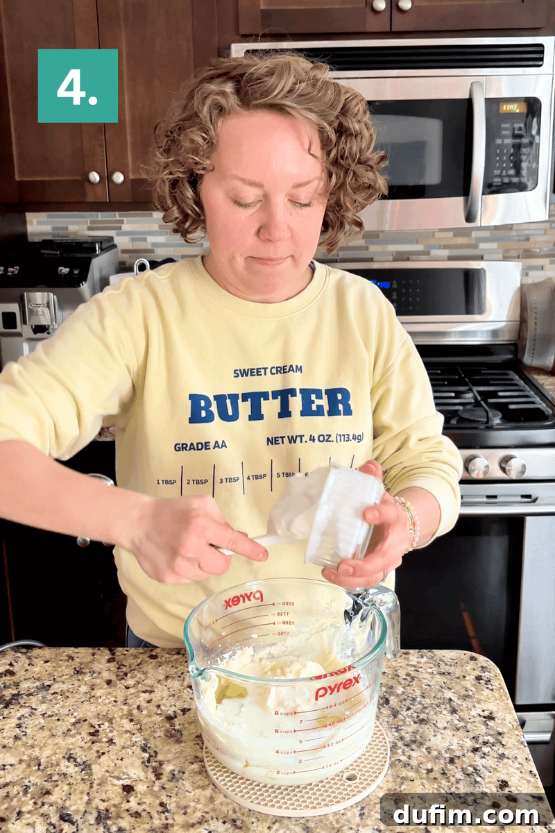 A person adds an ingredient from a small container into a glass measuring cup with a mixture inside, standing in a kitchen. A green square with the number 4 is in the upper left corner.