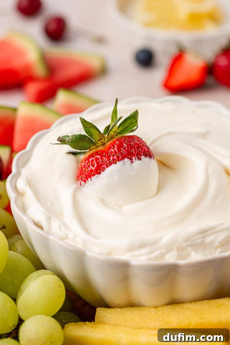 Cream Cheese Fruit Dip Recipe for Parties and Snacks 2 A white bowl filled with creamy fruit dip, topped with a whole strawberry, surrounded by fresh grapes, pineapple, and watermelon slices.