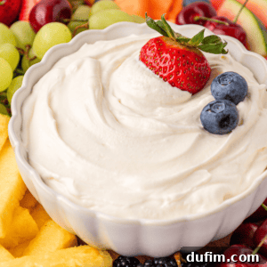 Cream Cheese Fruit Dip Recipe for Parties and Snacks 12 A white bowl filled with creamy fruit dip, topped with a strawberry and two blueberries, surrounded by fresh fruit pieces including pineapple, grapes, berries, and melon.