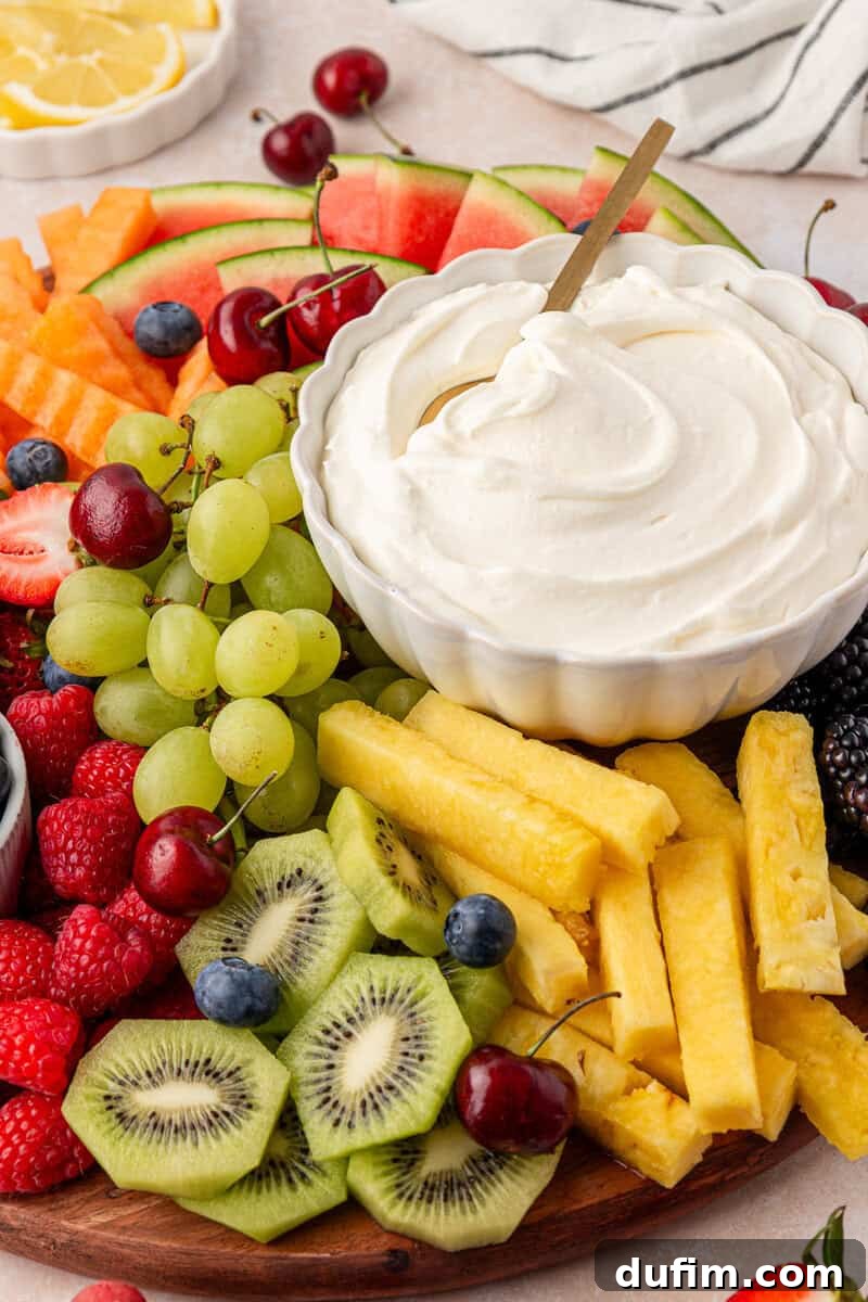 Cream Cheese Fruit Dip Recipe for Parties and Snacks 3 A platter of assorted fresh fruits including kiwi, pineapple, grapes, watermelon, strawberries, raspberries, blueberries, cherries, and blackberries, arranged around a bowl of creamy fruit dip with a spoon.
