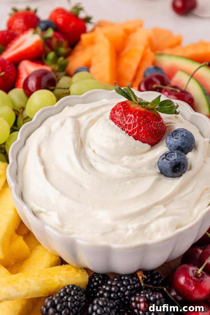 Cream Cheese Fruit Dip Recipe for Parties and Snacks 10 A white bowl of creamy fruit dip topped with a strawberry and two blueberries, surrounded by assorted fresh fruits.