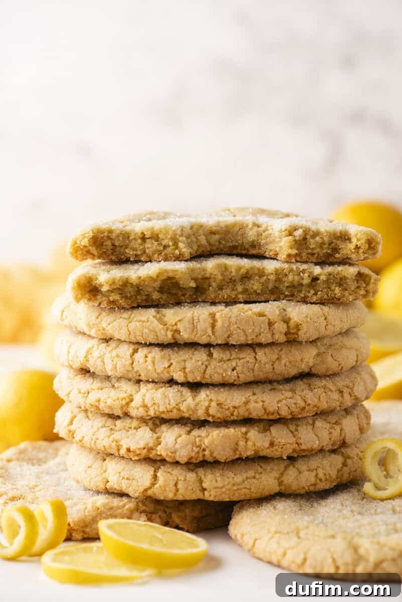 A stack of sugar-coated lemon cookies is shown, with the top cookie broken in half to reveal a soft, crumbly interior. Lemon slices and lemon zest curls are scattered around the cookies.
