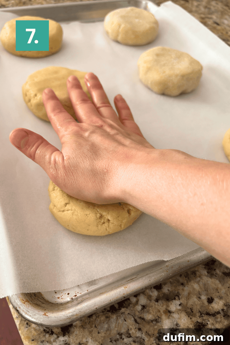 A hand presses down on a round ball of dough, flattening it on a parchment-lined baking sheet.