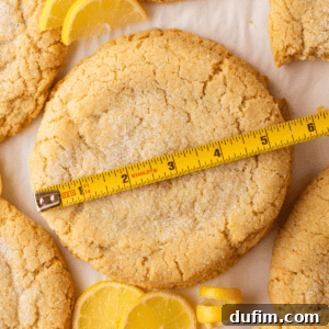 A large, round lemon sugar cookie topped with sugar is measured by a yellow tape measure. Lemon slices and other cookies are visible around it on a light surface.