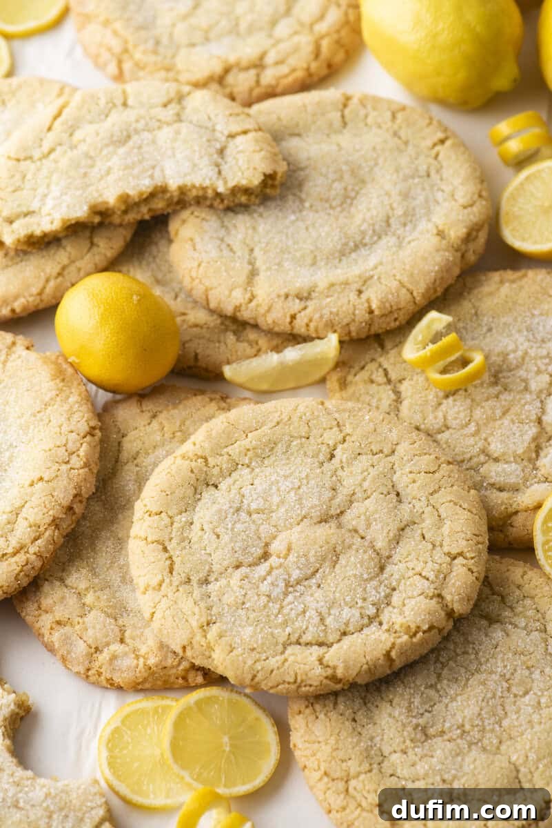 Large, round lemon sugar cookies are arranged on a surface with fresh whole and sliced lemons scattered around. One cookie is partially eaten, showing the soft, crumbly interior.