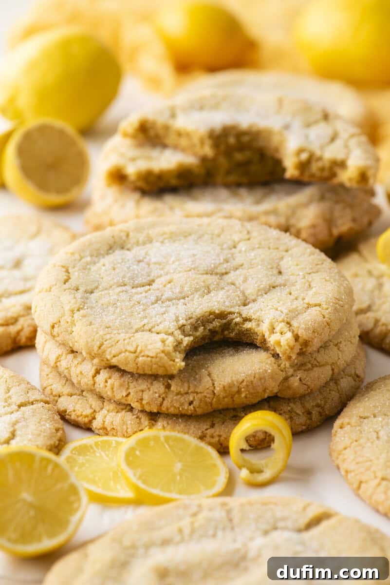 A stack of sugar-coated lemon cookies, with one cookie on top showing a bite taken out. Fresh lemon slices and whole lemons are scattered around the cookies.