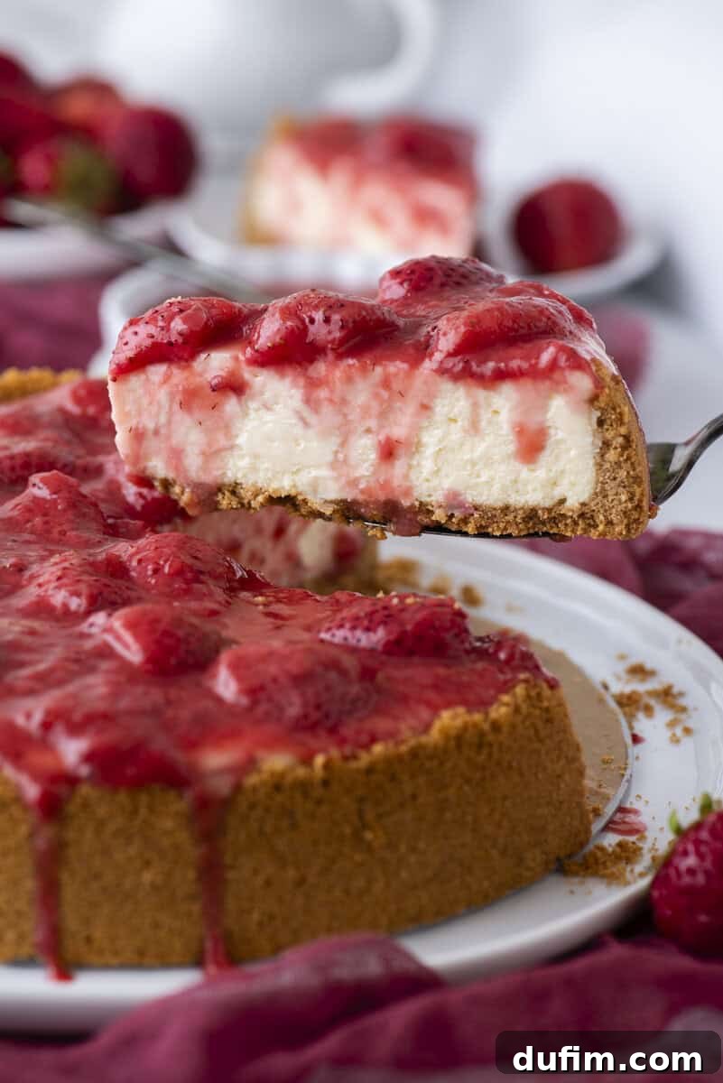 Strawberry Cheesecake Recipe with Fresh Strawberry Topping 2 A close-up of a creamy cheesecake topped with strawberry sauce and fresh strawberries. A slice is being lifted from the whole cake, revealing the thick graham cracker crust and smooth filling.
