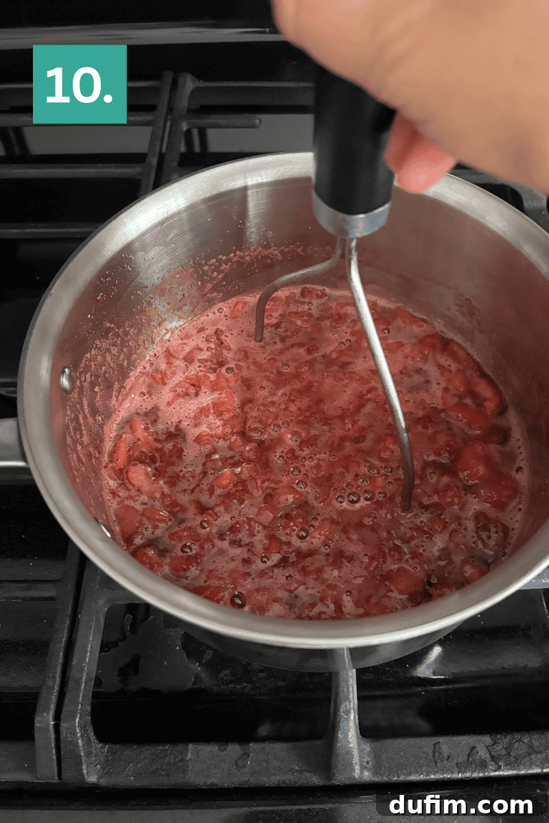 Strawberry Cheesecake Recipe with Fresh Strawberry Topping 13 A hand uses a potato masher to mash cooked strawberries in a pot on a stove.