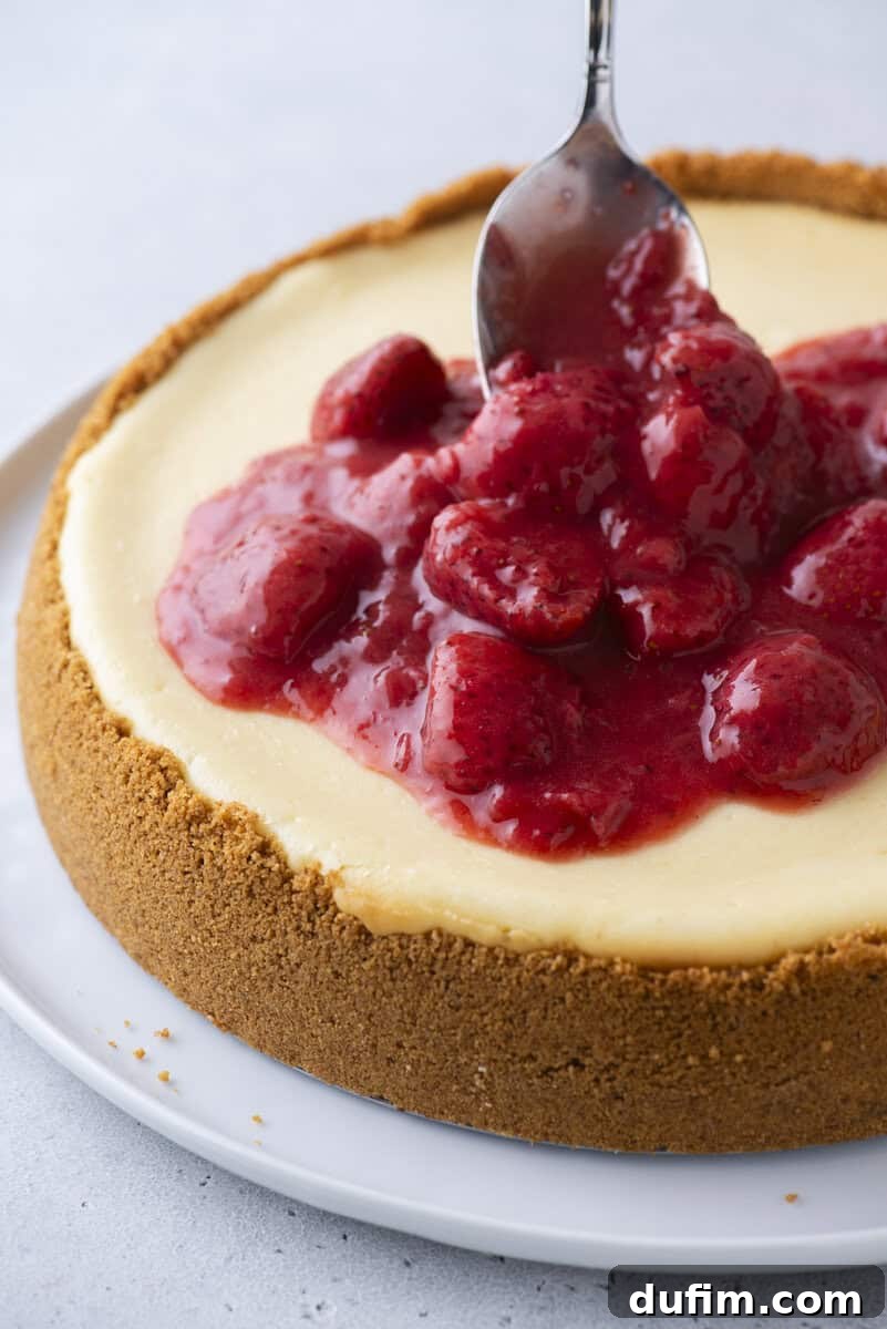 Strawberry Cheesecake Recipe with Fresh Strawberry Topping 16 A spoon spreads glossy strawberry topping over a creamy cheesecake with a thick graham cracker crust on a white plate.
