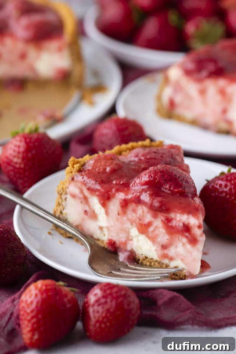 Strawberry Cheesecake Recipe with Fresh Strawberry Topping 19 A slice of strawberry cheesecake with a graham cracker crust sits on a white plate, topped with strawberry sauce and whole strawberries.